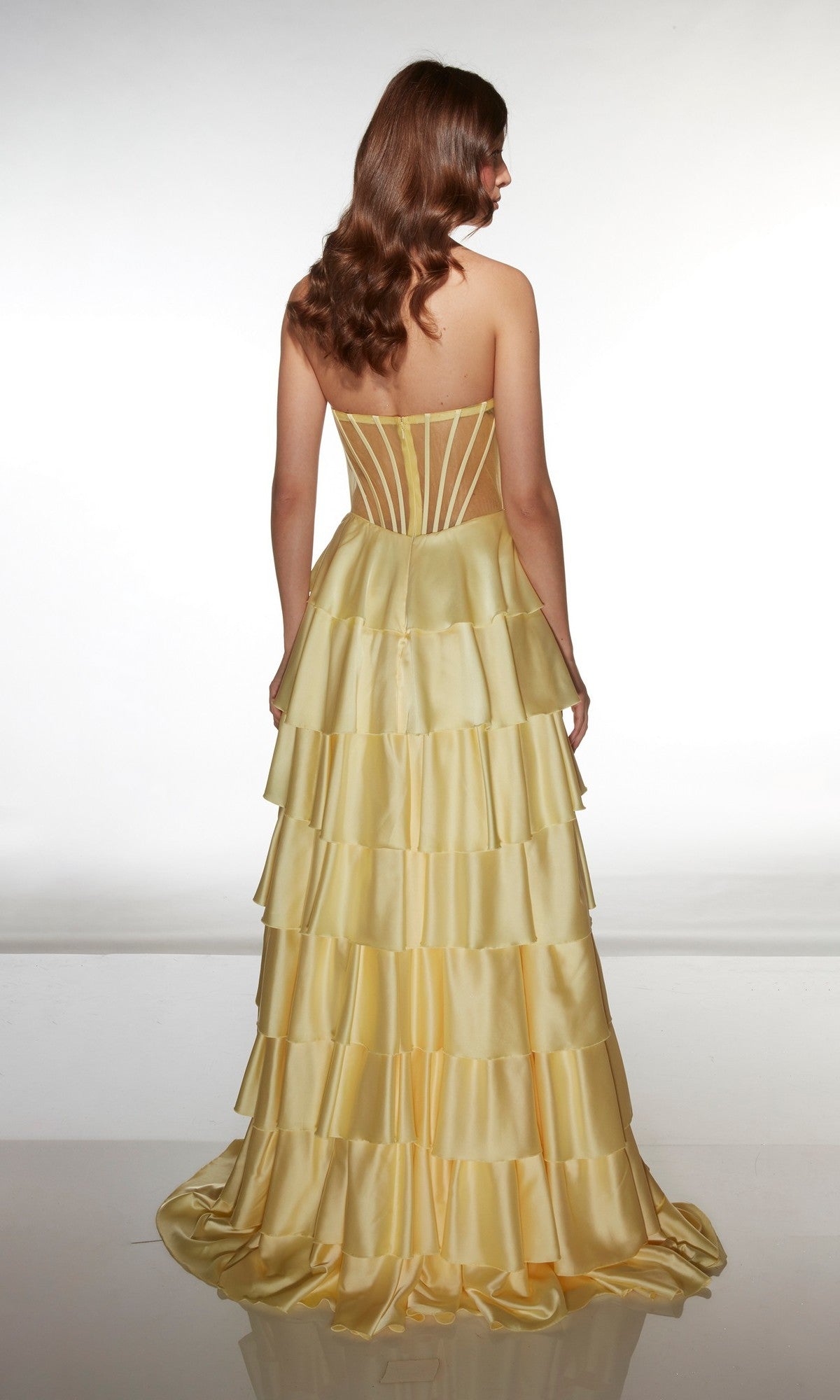 A woman with wavy brown hair stands facing away, wearing the Alyce 62056 long prom dress—a strapless satin gown with a sweetheart neckline and tiered ruffle A-line skirt in vibrant yellow.